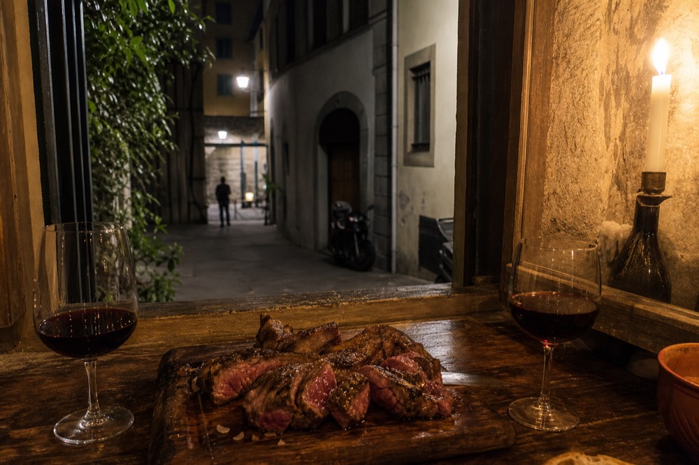 Glasses of red wine and a plate of sliced steak on a rustic table in a candlelit Florentine restaurant