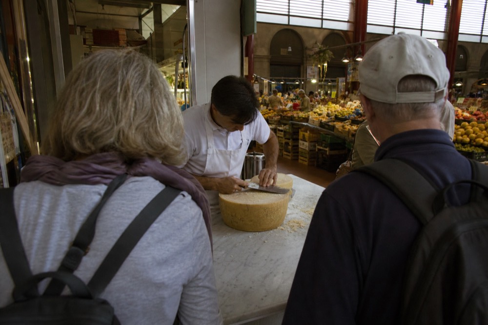 A food tour group standing around a market stall while a vendor slices cheese