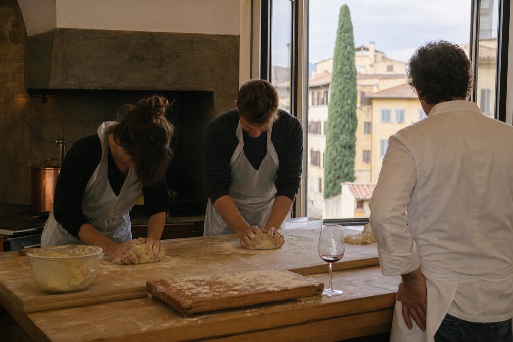 Two people knead pasta dough at a cooking station while a chef looks on