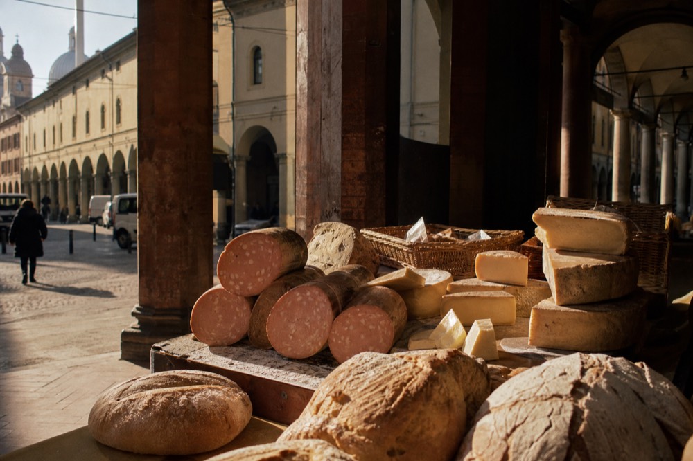 A market stall display of cured meats, cheeses and loaves of bread arranged in rows