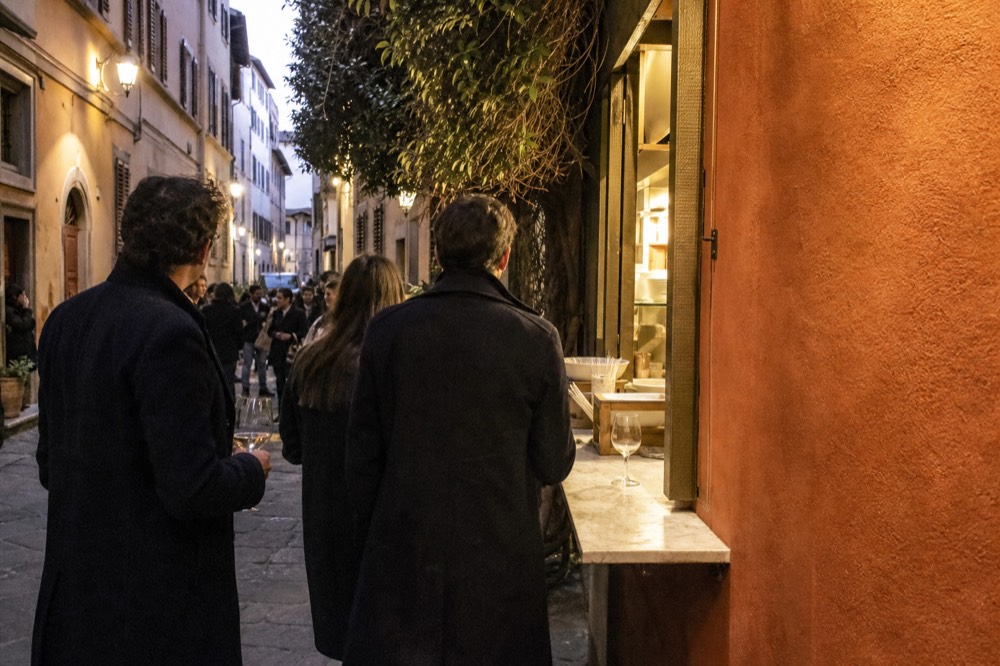 A small group of standing diners holding wine glasses near an open-air food counter in the early evening