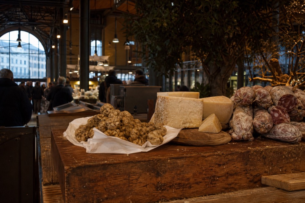 A wooden market stall with truffles, cheese wedges and cured meats under warm lighting