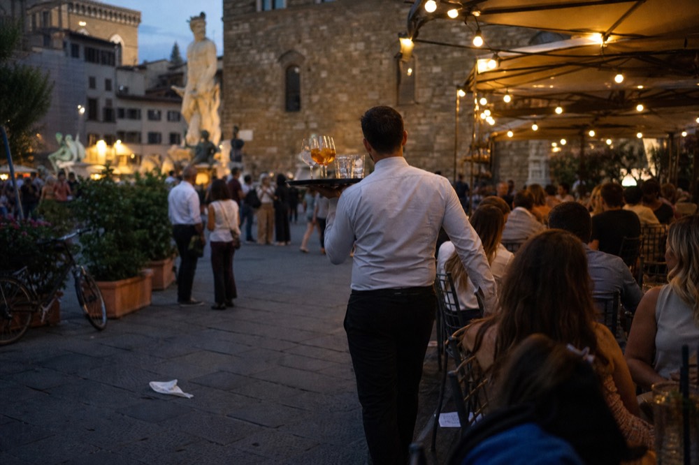 Waiter carrying a tray of drinks through a crowded evening restaurant terrace in Florence