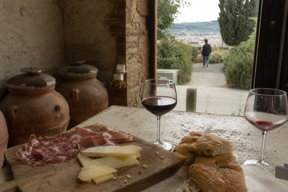 A close-up of a wooden board with slices of finocchiona, pecorino, prosciutto, and schiacciata, with a glass of red wine beside it