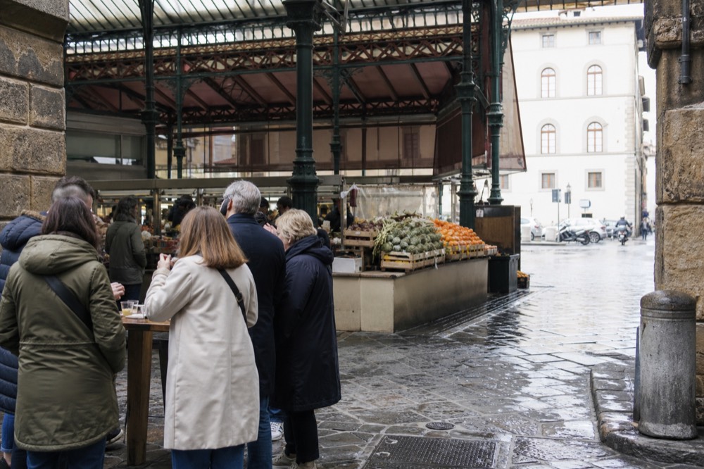 A food tour group standing at a high table inside a covered market, tasting olive oil from small cups, with produce stalls visible behind them