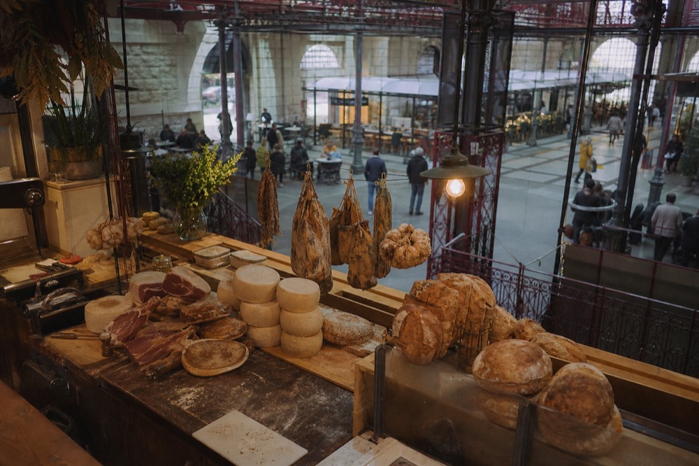 A wooden market stall display with cured meats, cheese wheels and loaves of bread
