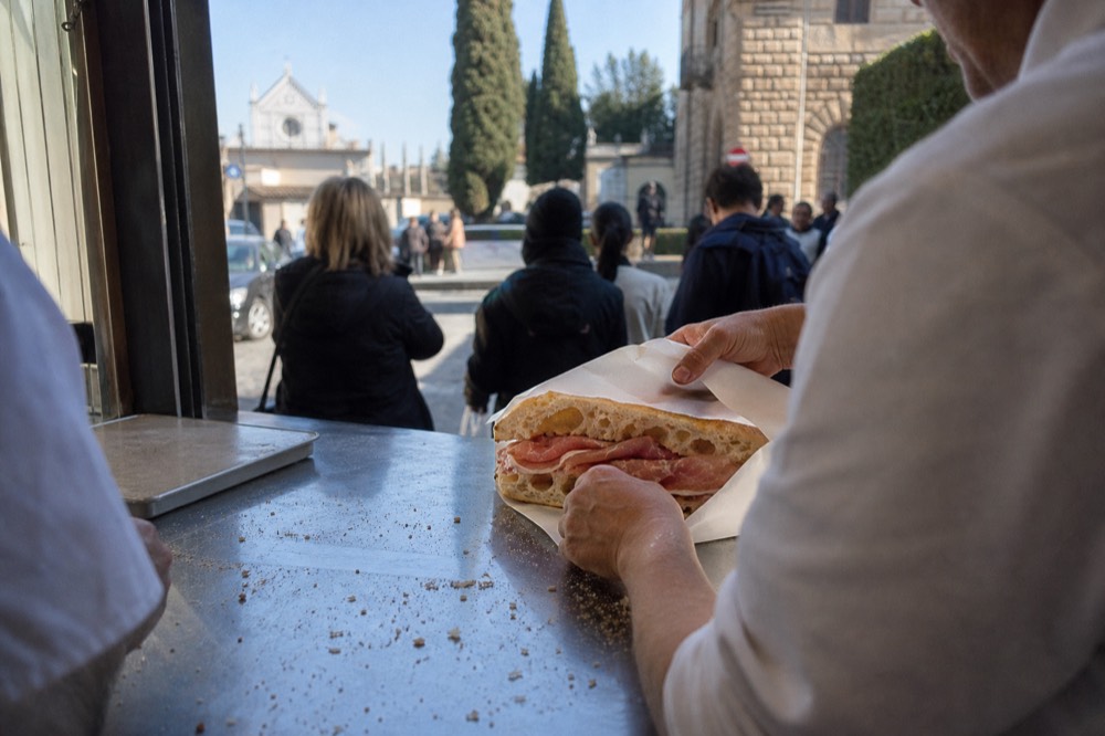 A schiacciata sandwich being wrapped in paper at a busy Florence bakery counter